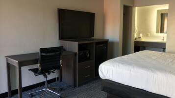 Habitación de hotel con cama, escritorio con silla, televisor, cómoda y baño con lavamanos y espejo.