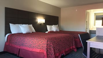 A hotel room with two beds, each with white pillows and red patterned bedspreads. There is a bedside table with a lamp and a small desk with a chair.