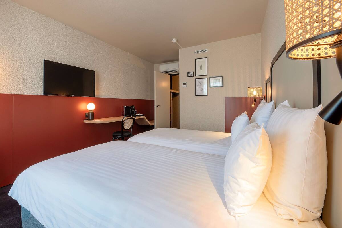 A hotel room with a large bed, a flat-screen TV mounted on the wall, a desk with a chair, and a bedside table with a lamp.