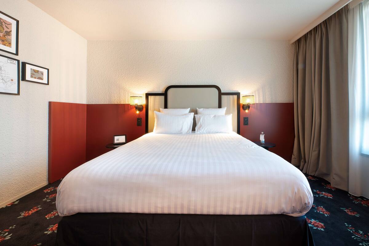 A hotel room with a large bed, two bedside tables, wall-mounted lamps, and a window with curtains.