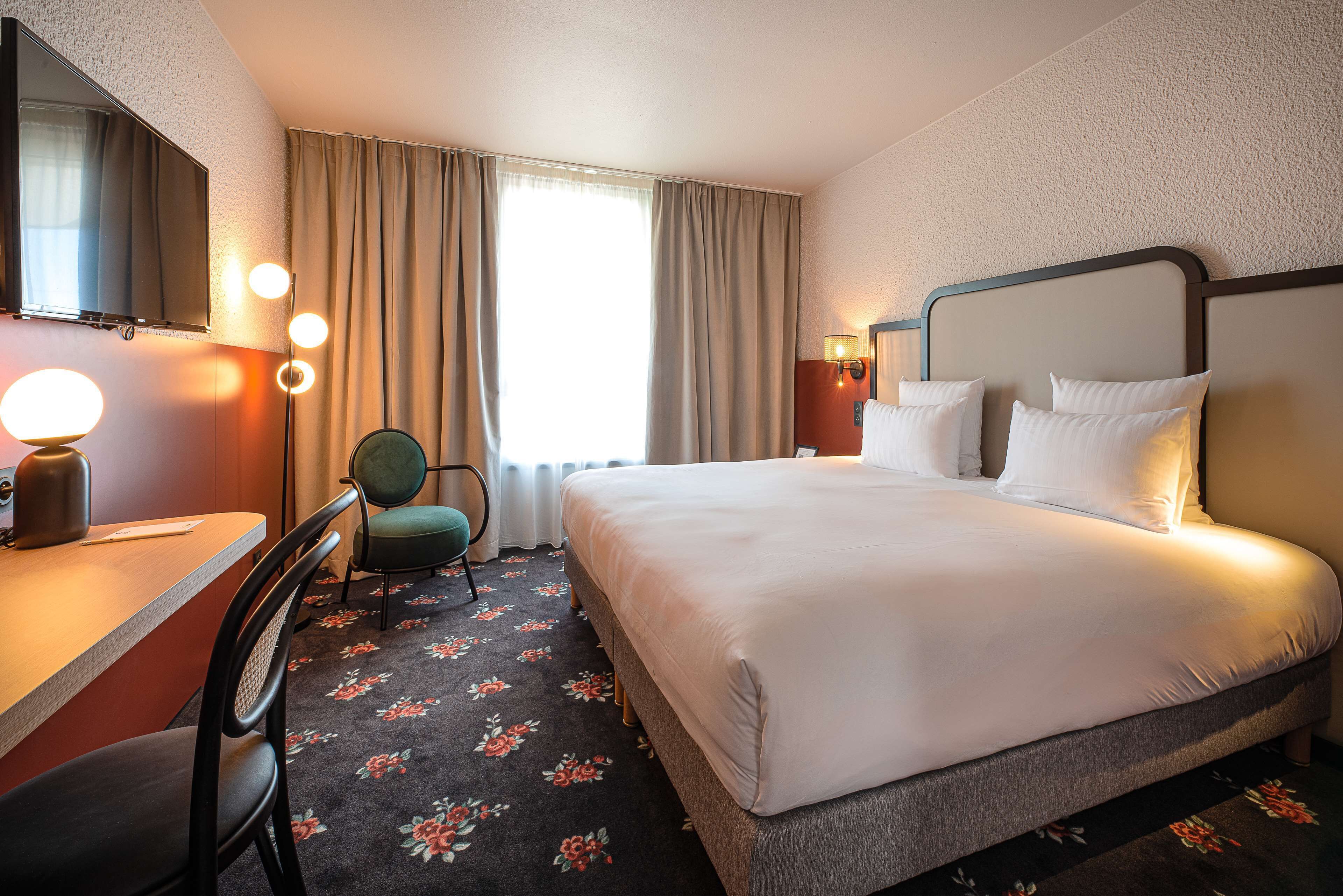 Ein Hotelzimmer mit Bett, Schreibtisch, Stuhl, Fernseher und einem Fenster mit Vorhängen.