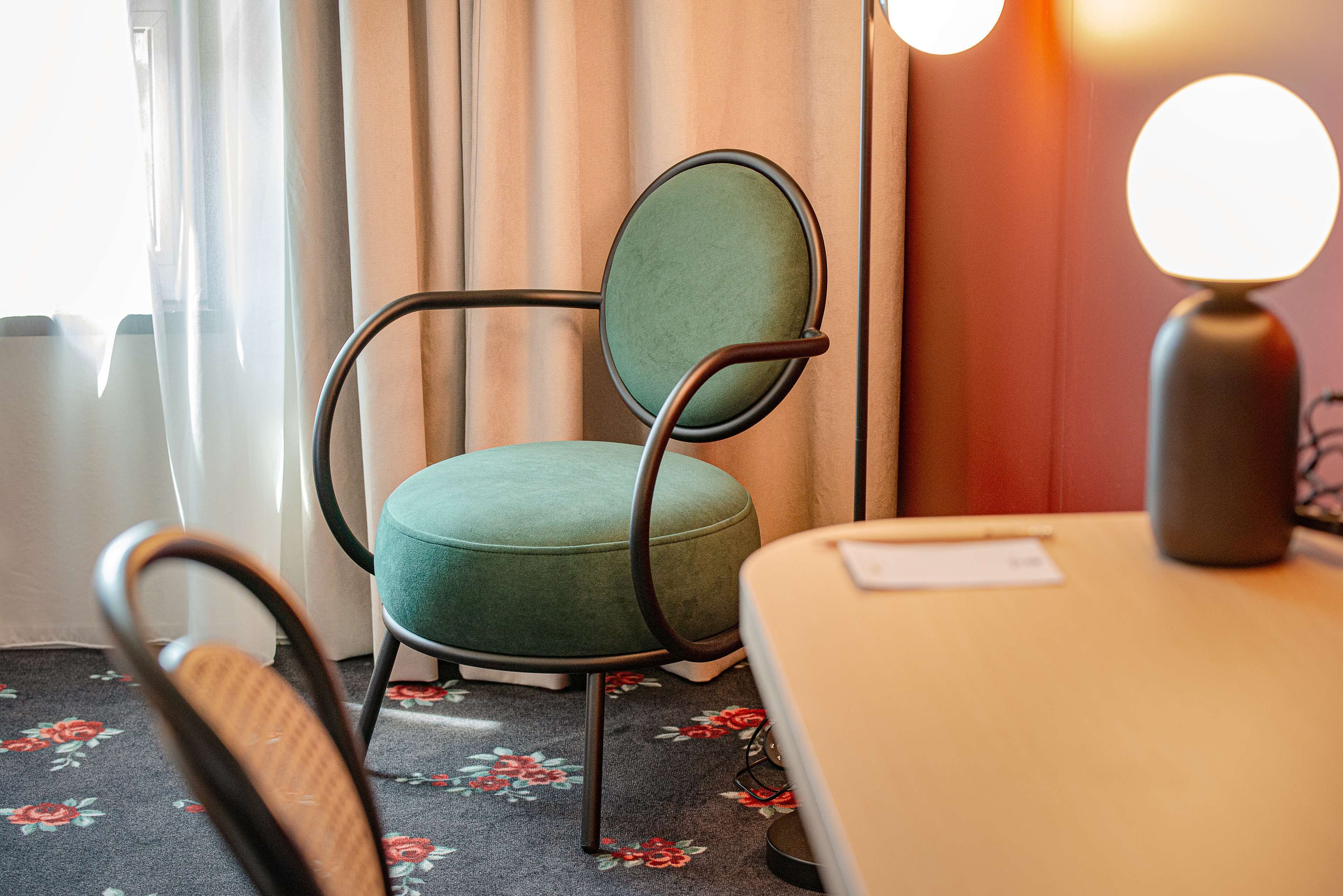 A room with a green upholstered chair, a wooden chair, a table with a lamp, and a floral carpet.