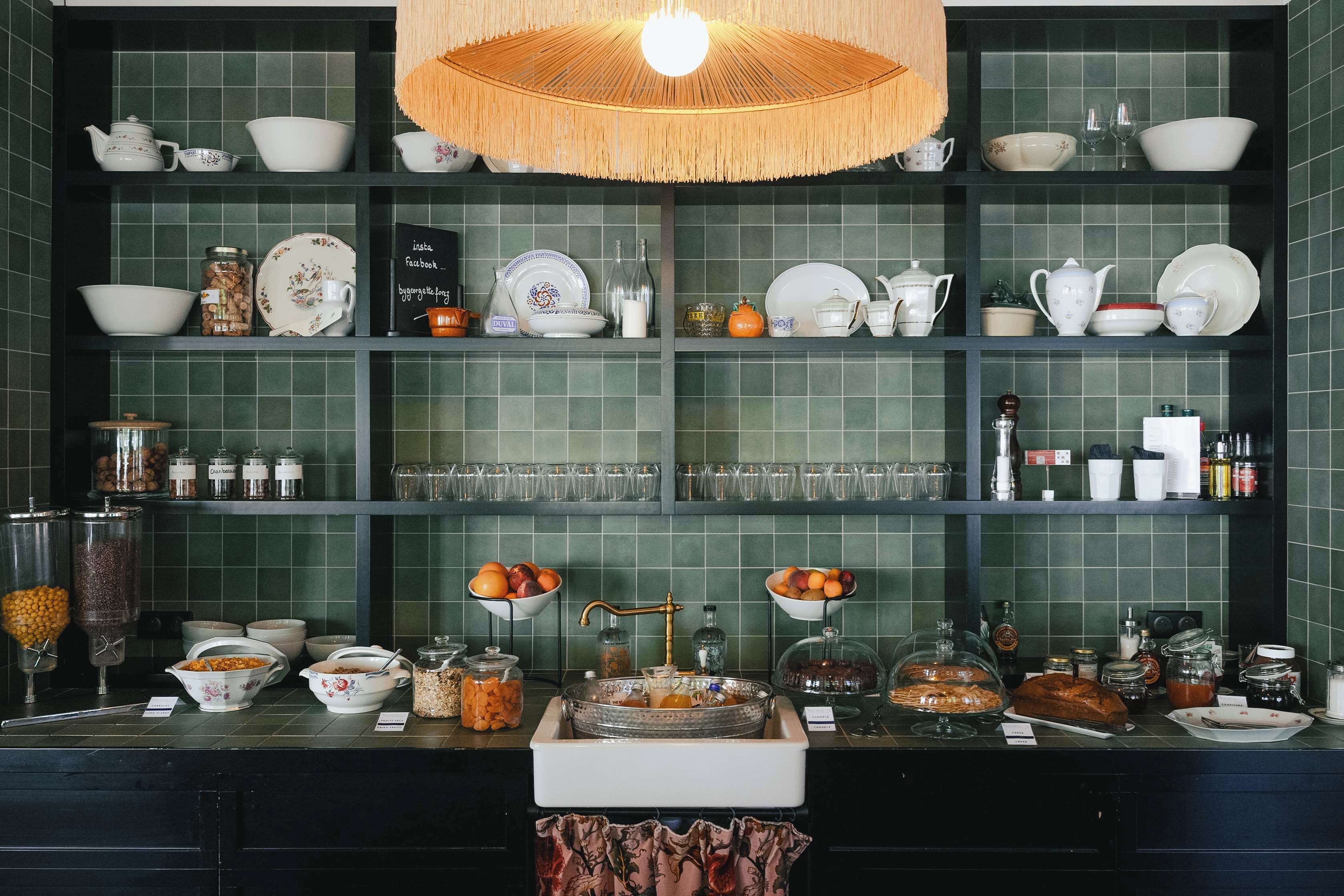 Una cocina con paredes de azulejos verdes, estantes con platos y frascos, y una isla central con comida y bebidas.
