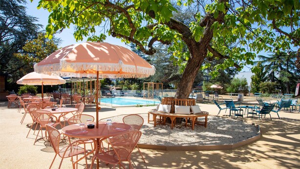 Ein Außenbereich mit Tischen, Stühlen und Sonnenschirmen, einem Pool und einem Baum, der Schatten spendet.