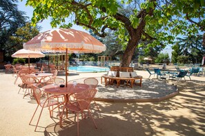 An outdoor seating area with tables and chairs under umbrellas, a pool, and a tree providing shade. - Best Western Saint-Etienne Porte du Forez (Andrezieux-Boutheon)