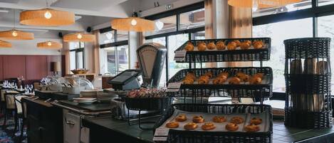 Um balcão de pastelaria com diversos produtos de panificação expostos em cestos de vime, uma máquina de café e um registo de caixa.
