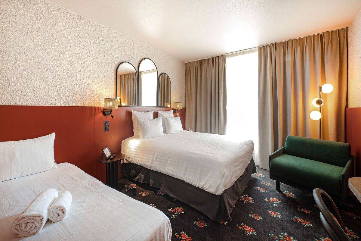 A hotel room with two beds, a green armchair, a mirror, and a nightstand with a lamp.