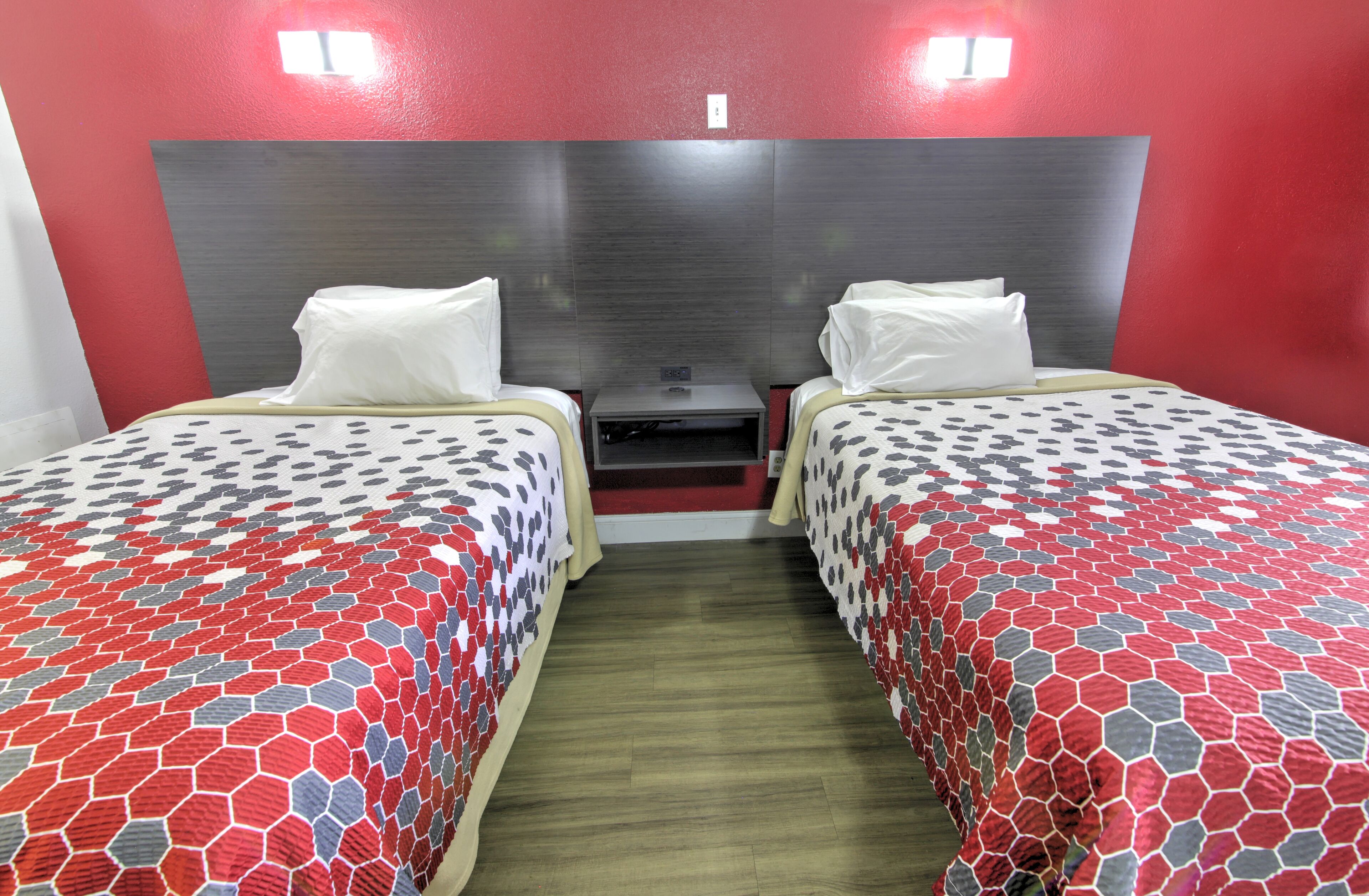 Quarto de hotel com duas camas, cada uma com colcha estampada, piso de madeira e parede em destaque na cor vermelha.