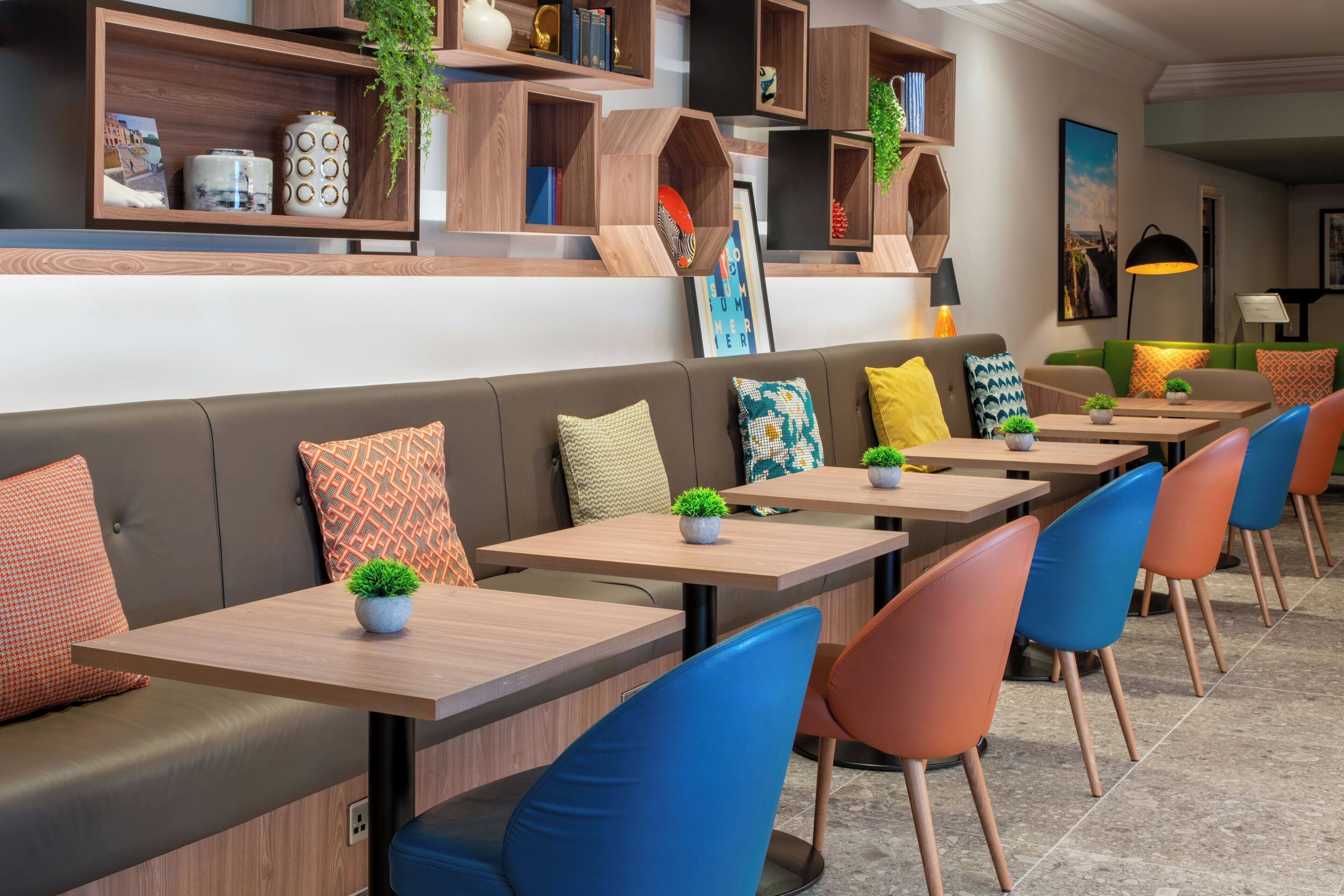 Un restaurante moderno con mesas de madera y sillas coloridas, un sofá con cojines estampados, y estanterías con plantas y libros.