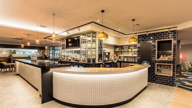 A modern bar area with a curved counter, overhead pendant lighting, and a tiled backsplash. - Holiday Inn Bristol-Filton by IHG (Bristol)