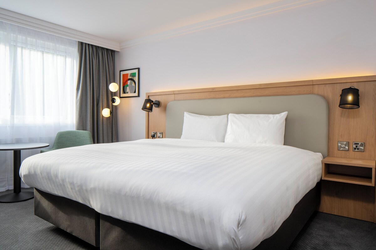 Een hotelkamer met een groot bed, een tafeltje, een stoel en een raam met gordijnen.