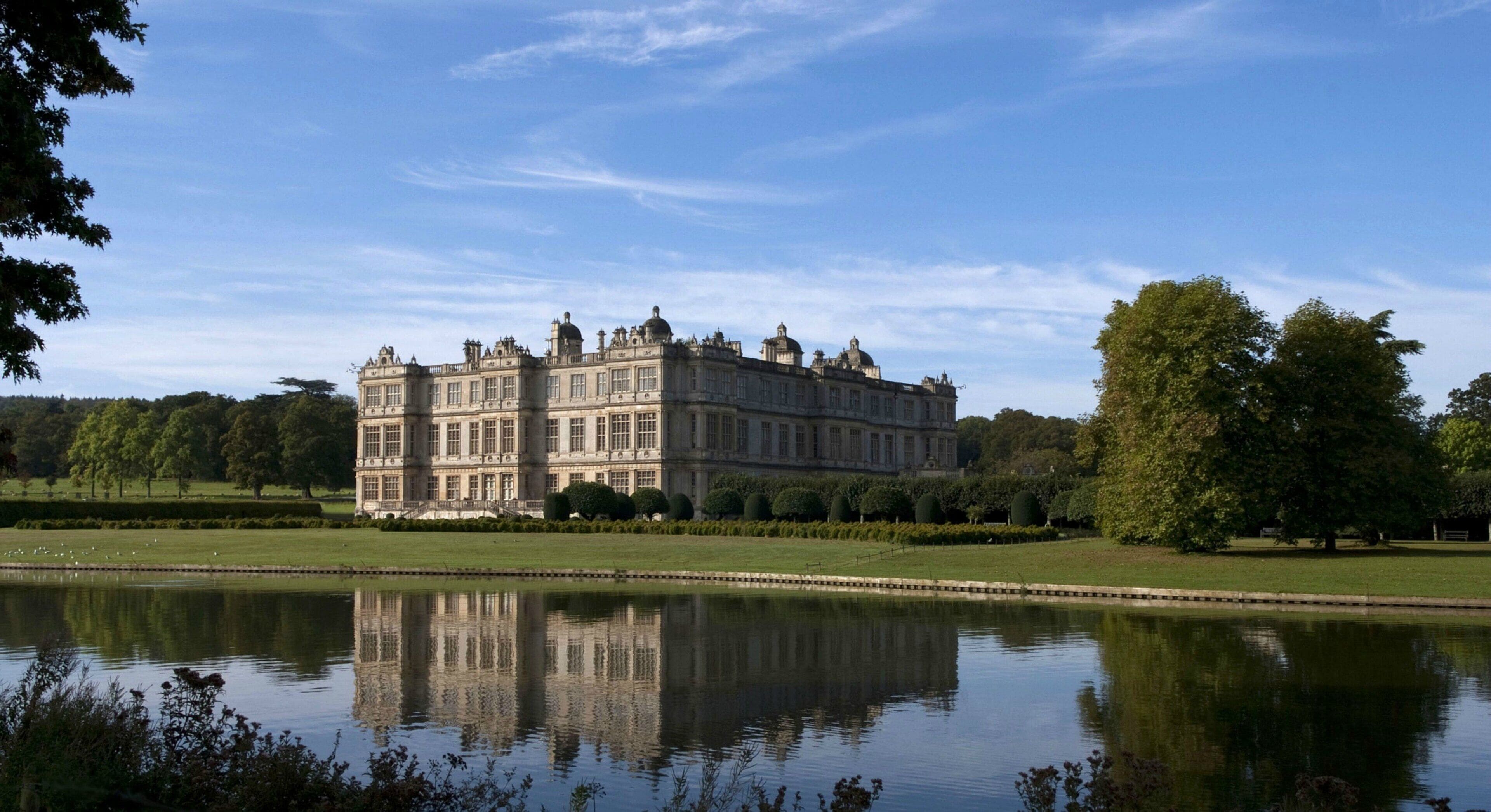 A large, historic building with a symmetrical facade, a reflection in the water, and a well-maintained lawn.