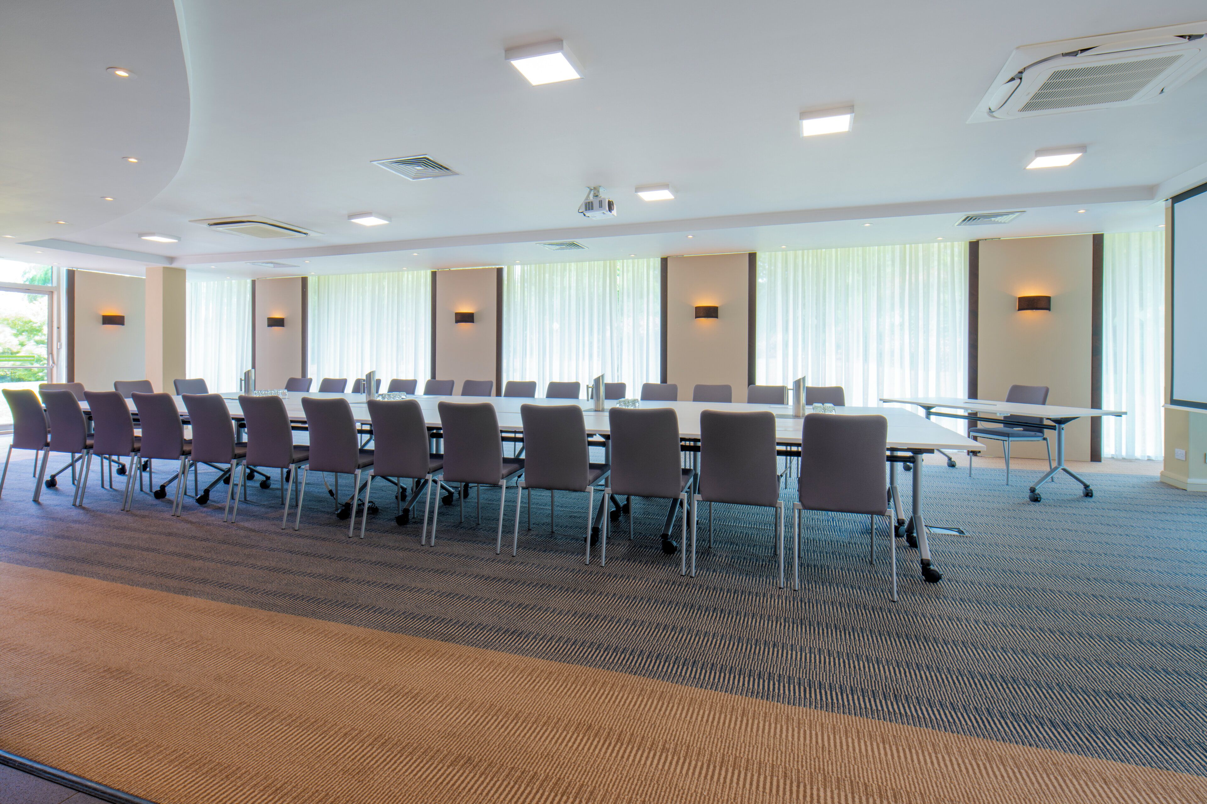 A conference room with a long table and chairs, large windows with blinds, and a carpeted floor.
