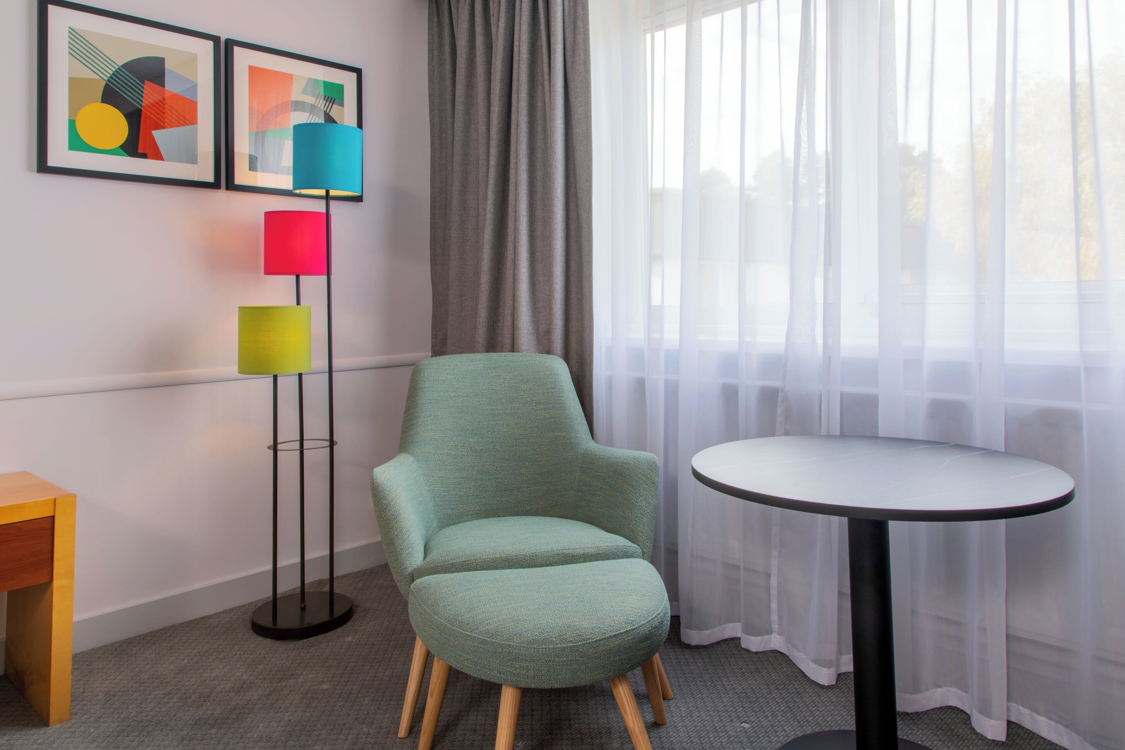 Een kamer met een groene fauteuil, een ronde tafel, een vloerlamp met meerdere gekleurde lampenkappen, en twee abstracte schilderijen aan de muur.