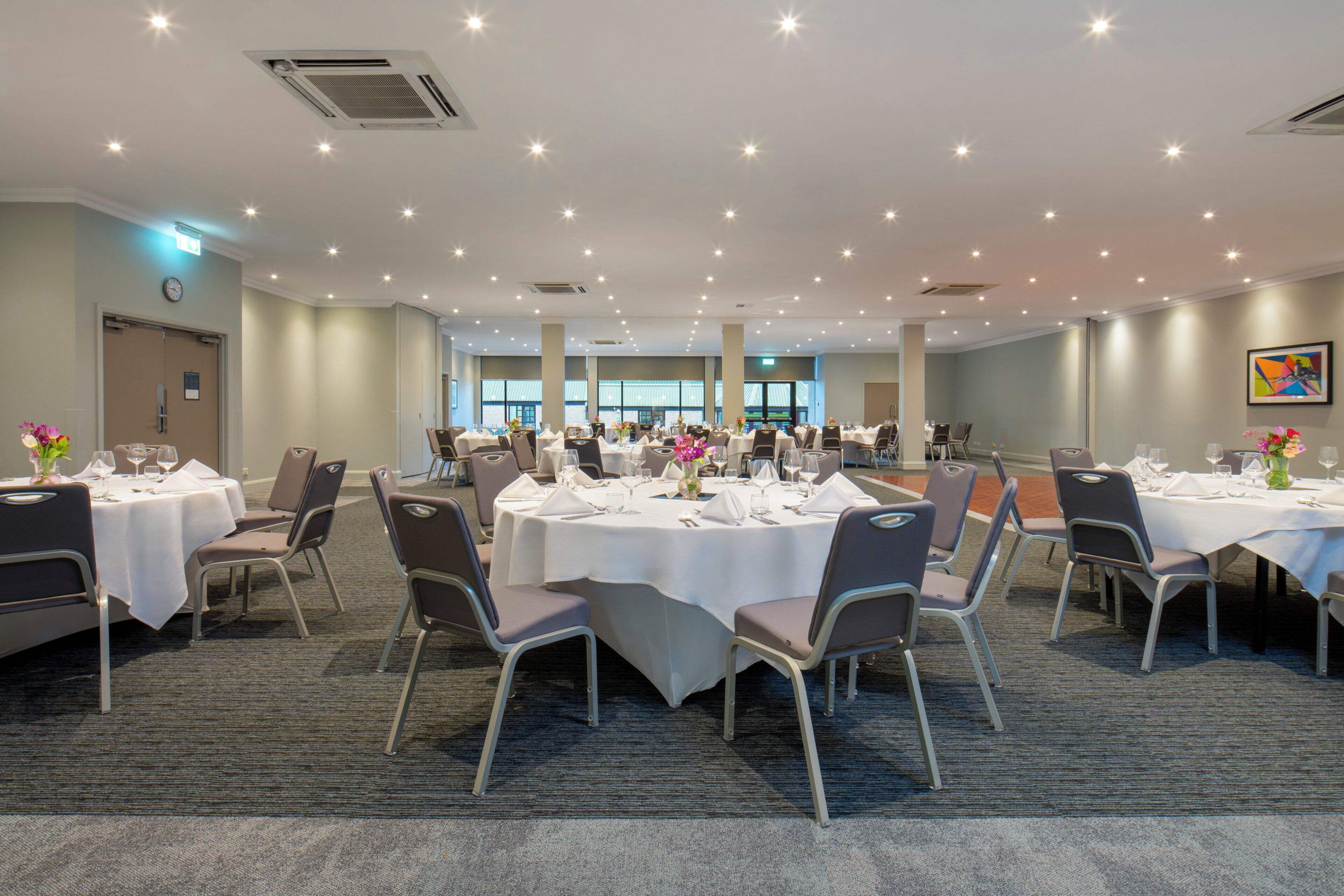 A large conference room with multiple tables and chairs, equipped with ceiling lights and air conditioning units.