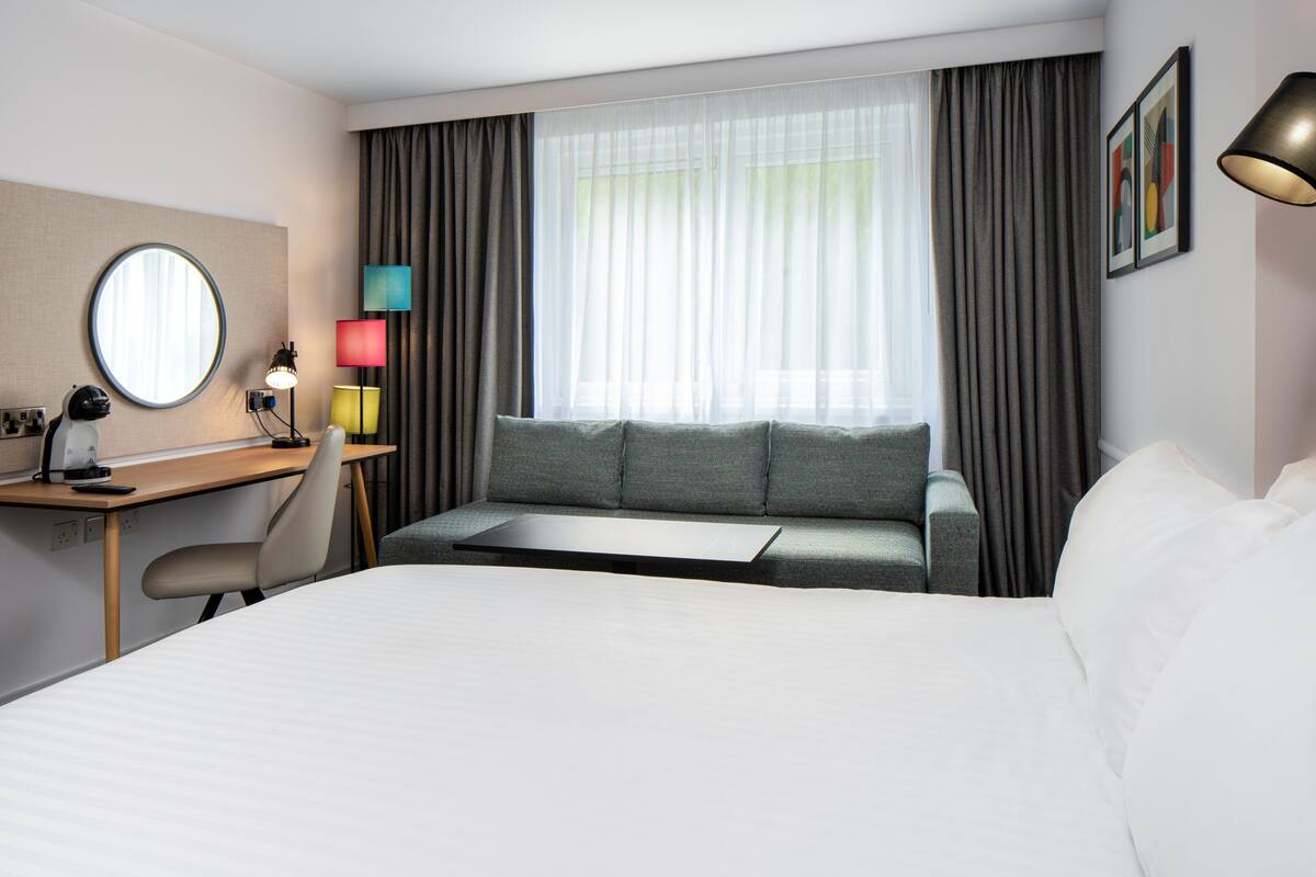 A hotel room with a bed, a desk with a chair, a sofa, a window with curtains, and a lamp.