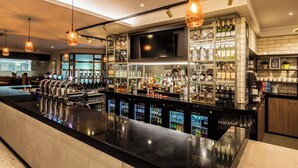 A bar area with a long counter, multiple beverage dispensers, and shelves stocked with various bottles and glasses. - Holiday Inn Bristol-Filton by IHG (Bristol)