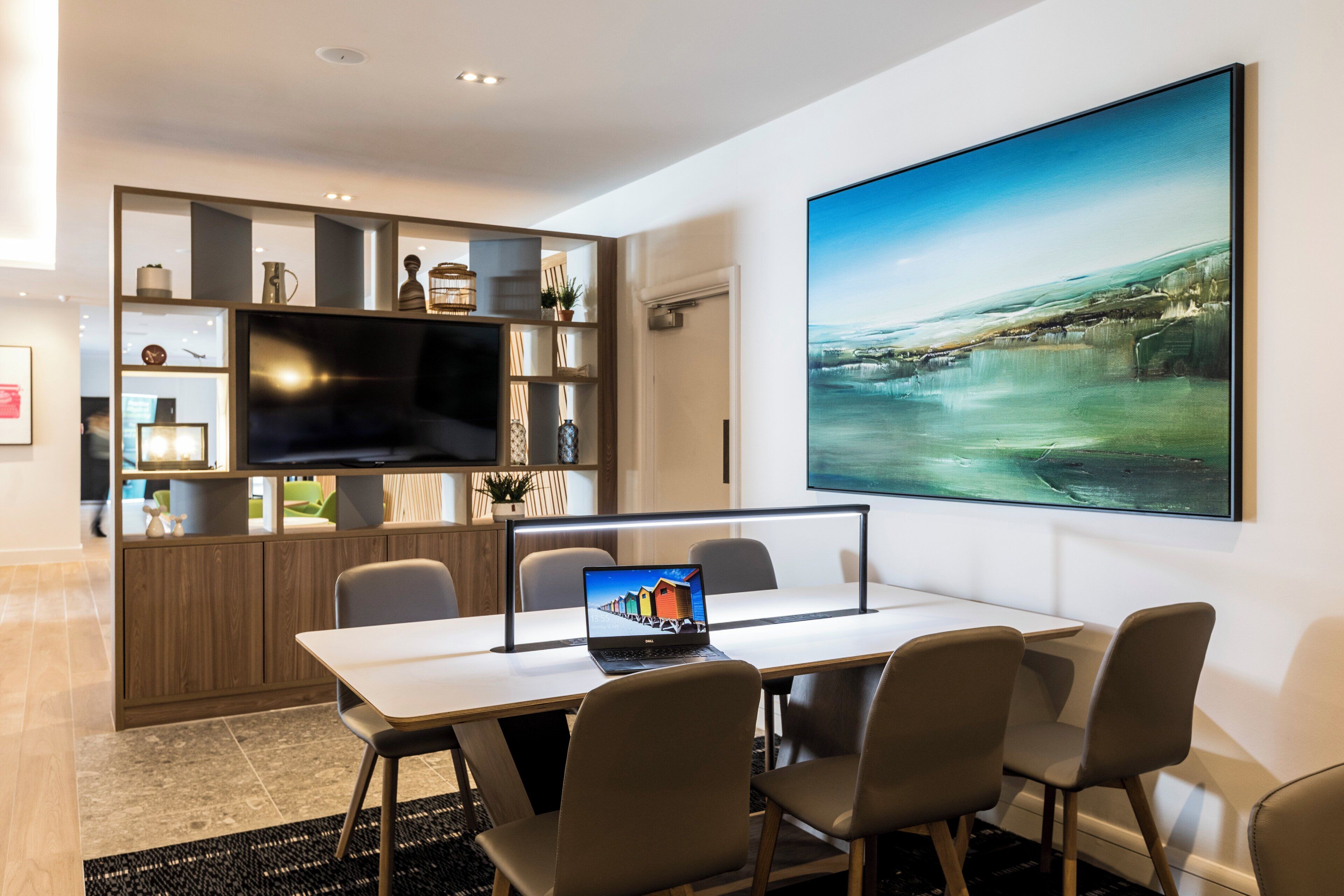 Una sala de reuniones moderna con un televisor de pantalla plana grande, una laptop Dell y un cuadro de paisaje de gran tamaño en la pared.