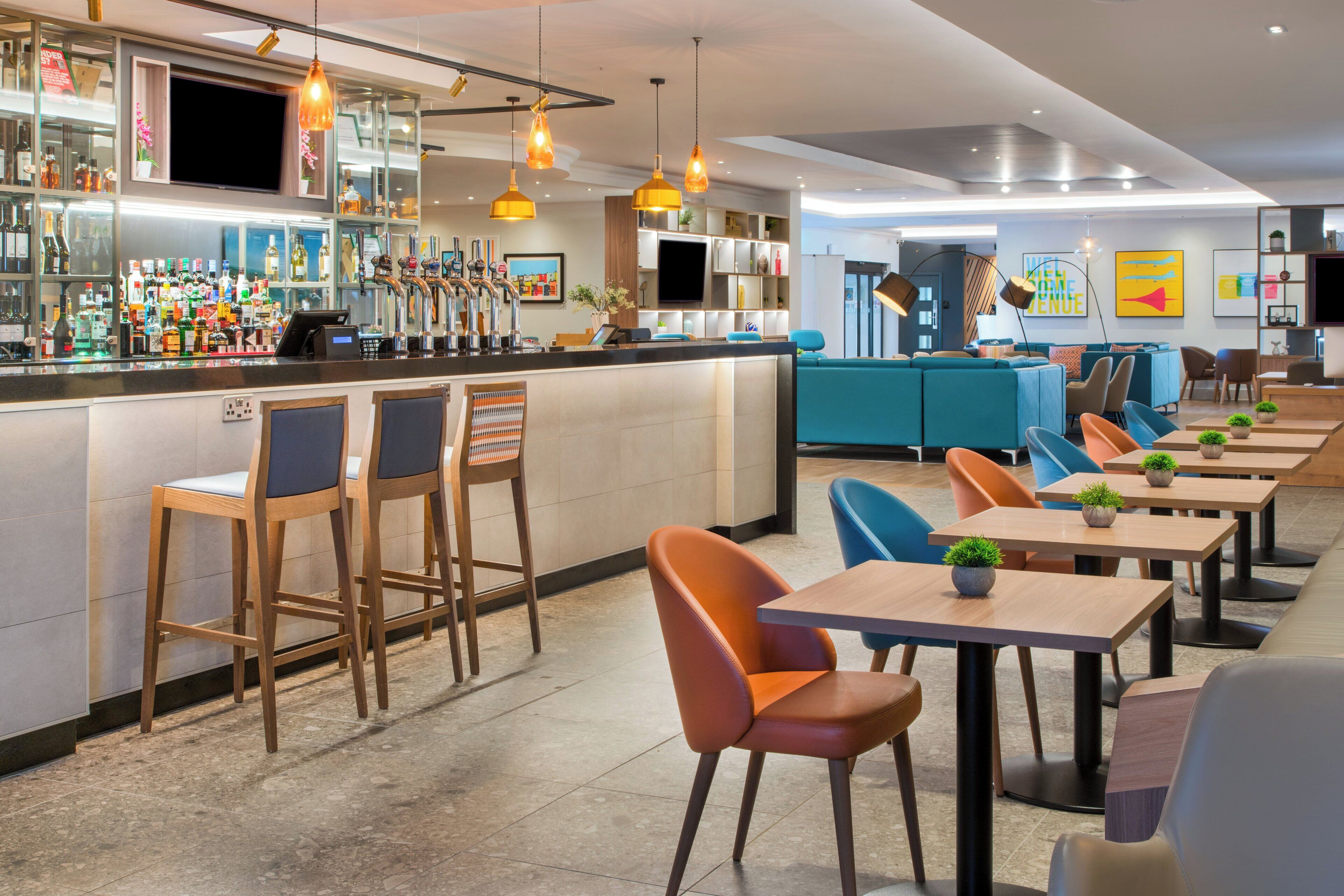 A modern bar area with a long counter, bar stools, and a variety of bottles and glasses. There are tables and chairs arranged for dining in the background.