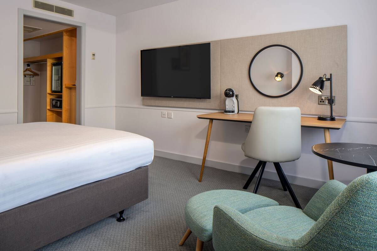 A hotel room with a bed, a desk with a computer, a chair, a round mirror, and a TV.