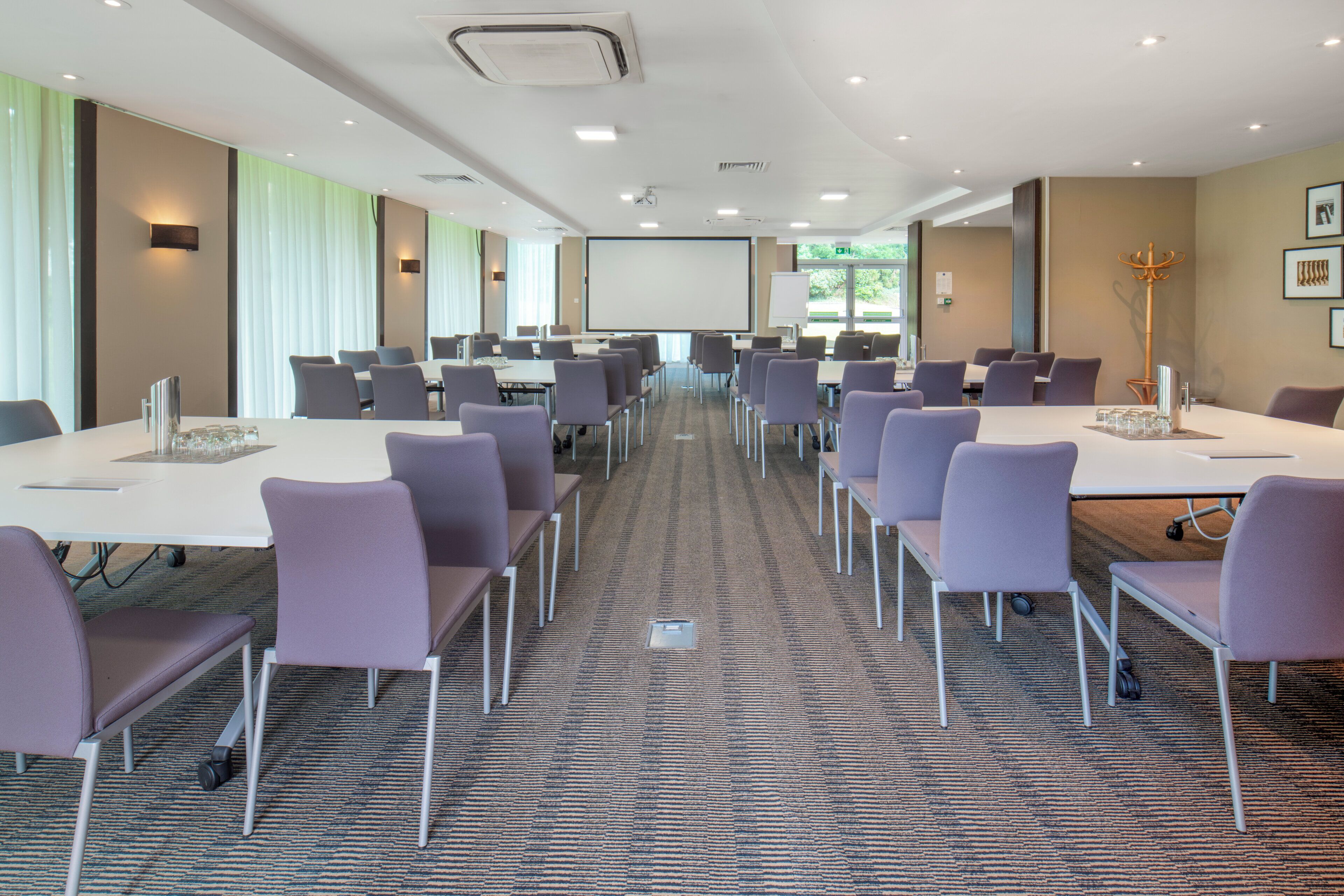 Un salón de conferencias con varias mesas y sillas, una pantalla de proyector y un gran ventanal con persianas.