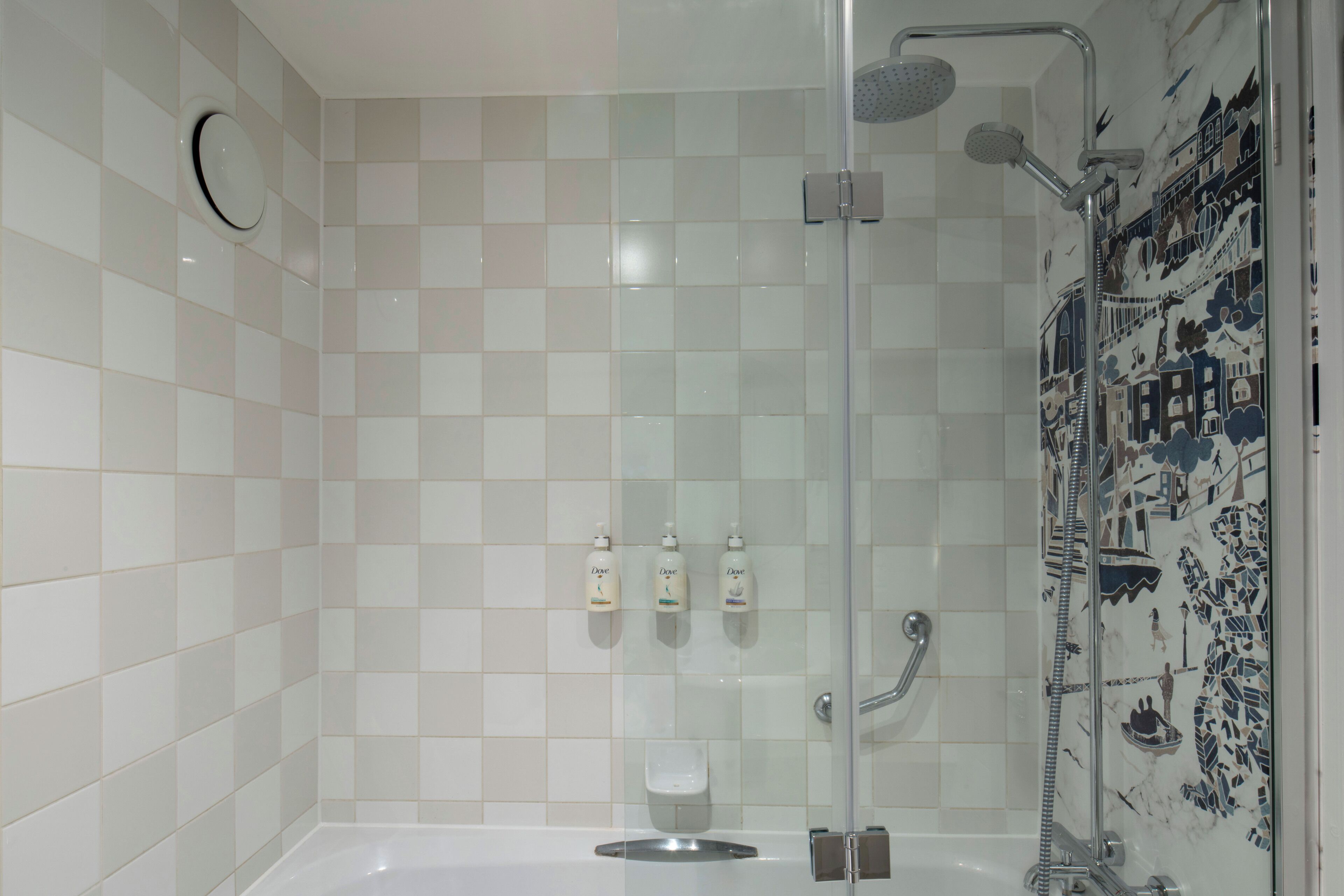 Un área de ducha moderna con puerta de cristal, cabezal de ducha manual y cabezal de ducha tipo lluvia. Hay tres jabones Dove en la pared.