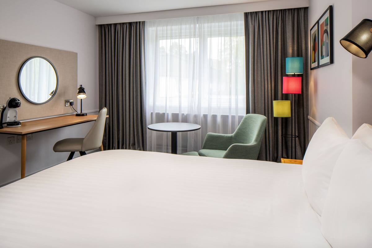 A hotel room with a bed, a desk, a chair, a round table, and a lamp.