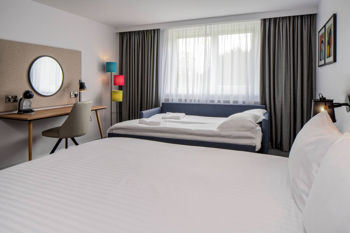 A hotel room with a bed, a desk, a chair, a round mirror, a lamp, a window with curtains, and a wall-mounted picture.