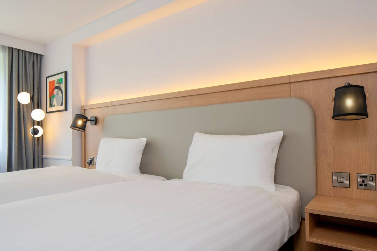 A hotel room with two beds, a wooden headboard, bedside lamps, a wall-mounted power outlet, and a framed picture.