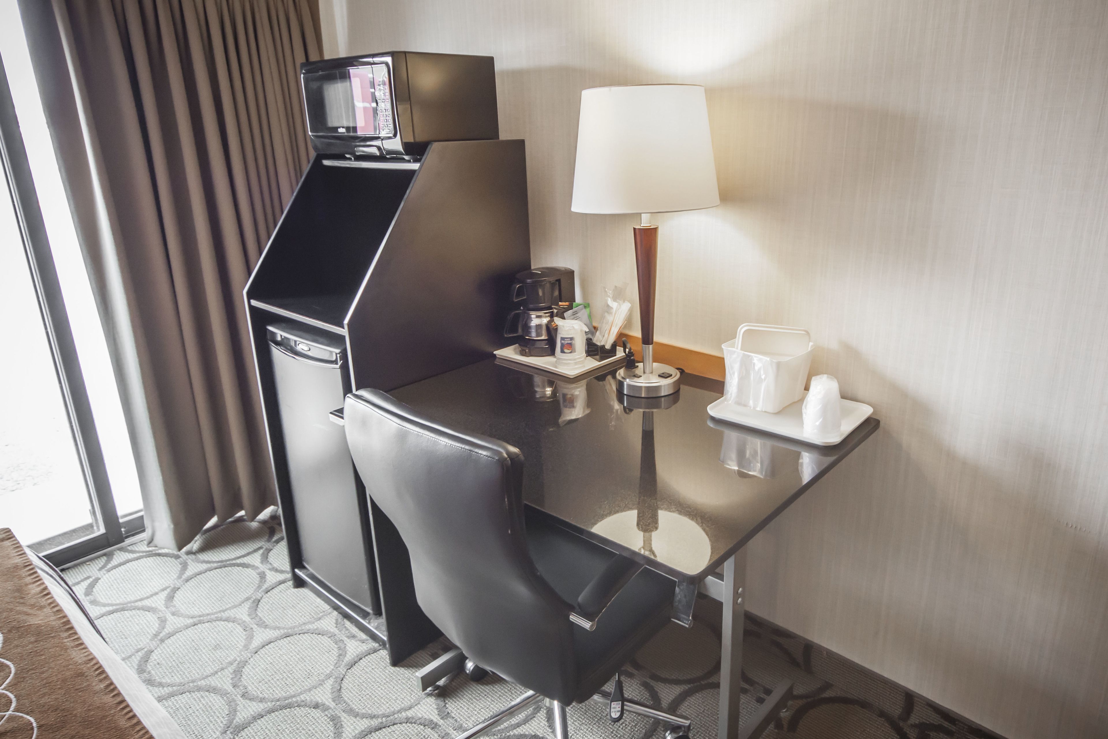 A hotel room with a compact desk, a coffee maker, a lamp, and a small table with cups and a bottle.