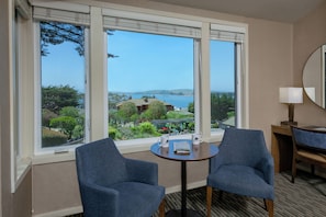 A hotel room with two blue armchairs, a small round table with a book and two mugs, and a view of a coastal landscape through large windows.