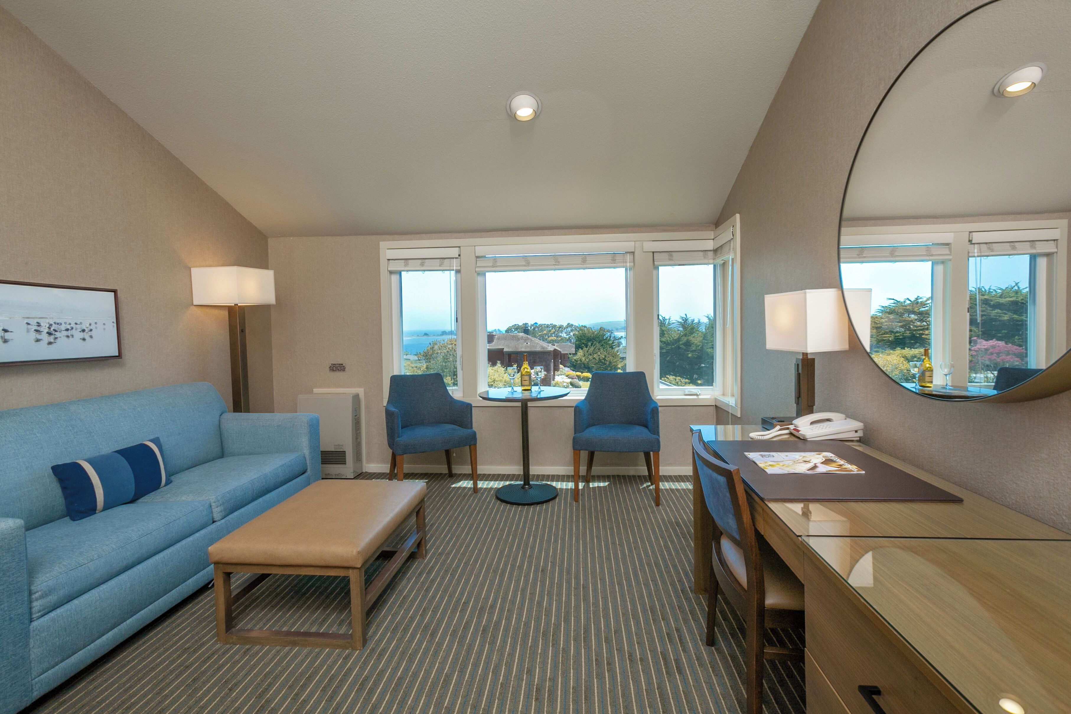 Une chambre d’hôtel avec un canapé bleu, deux fauteuils, une petite table, un bureau et un grand miroir. Il y a des fenêtres donnant sur l’extérieur.