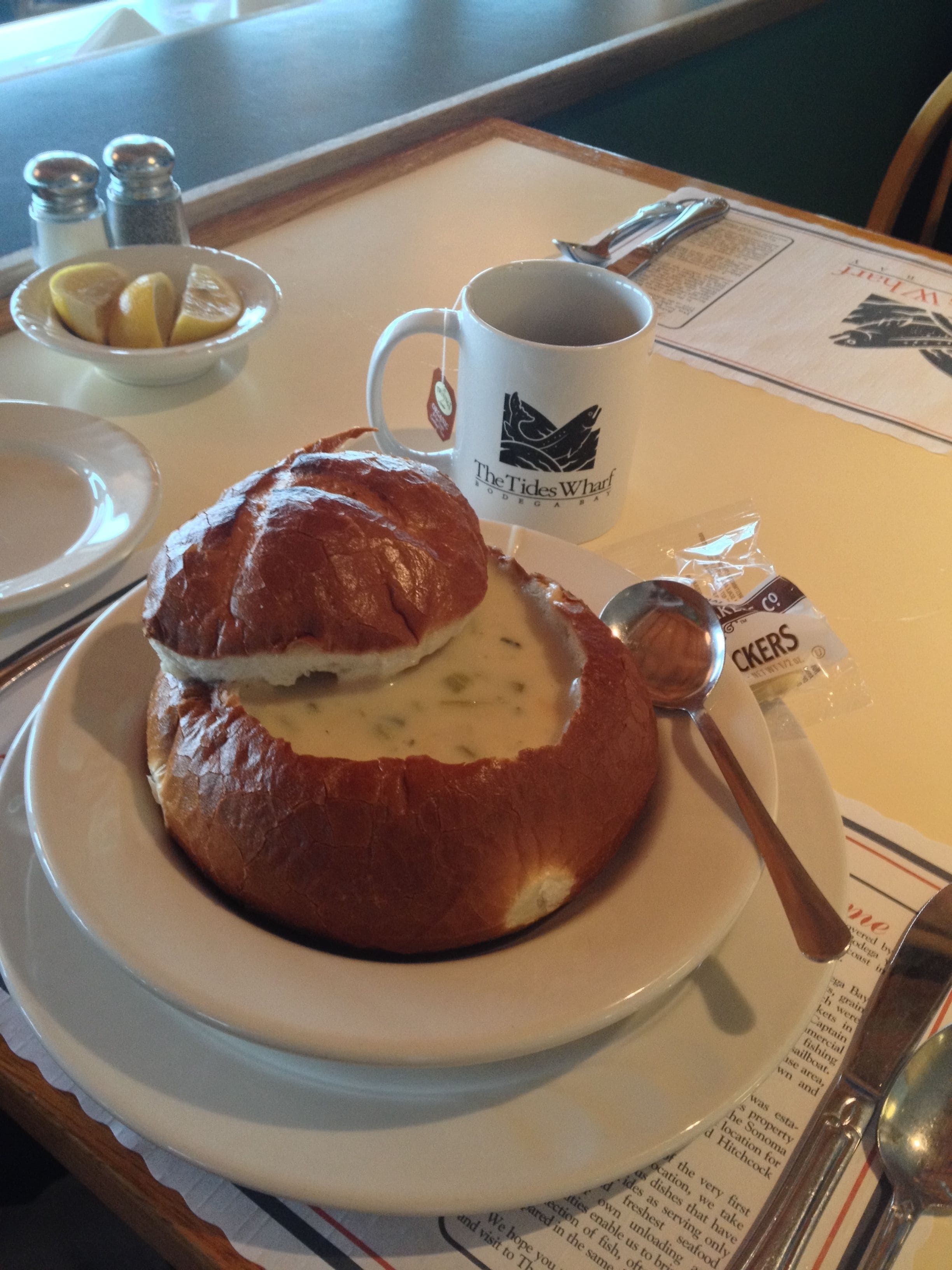 Un sándwich en un pan sobre un plato, una taza, saleros y pimenteros, un gajo de limón y una cuchara sobre una mesa.