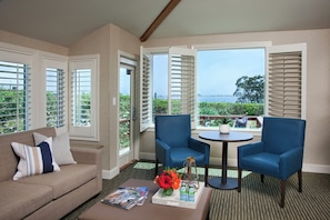 A room with a beige sofa, two blue armchairs, a small round table with a tray of drinks and flowers, and large windows with white shutters.