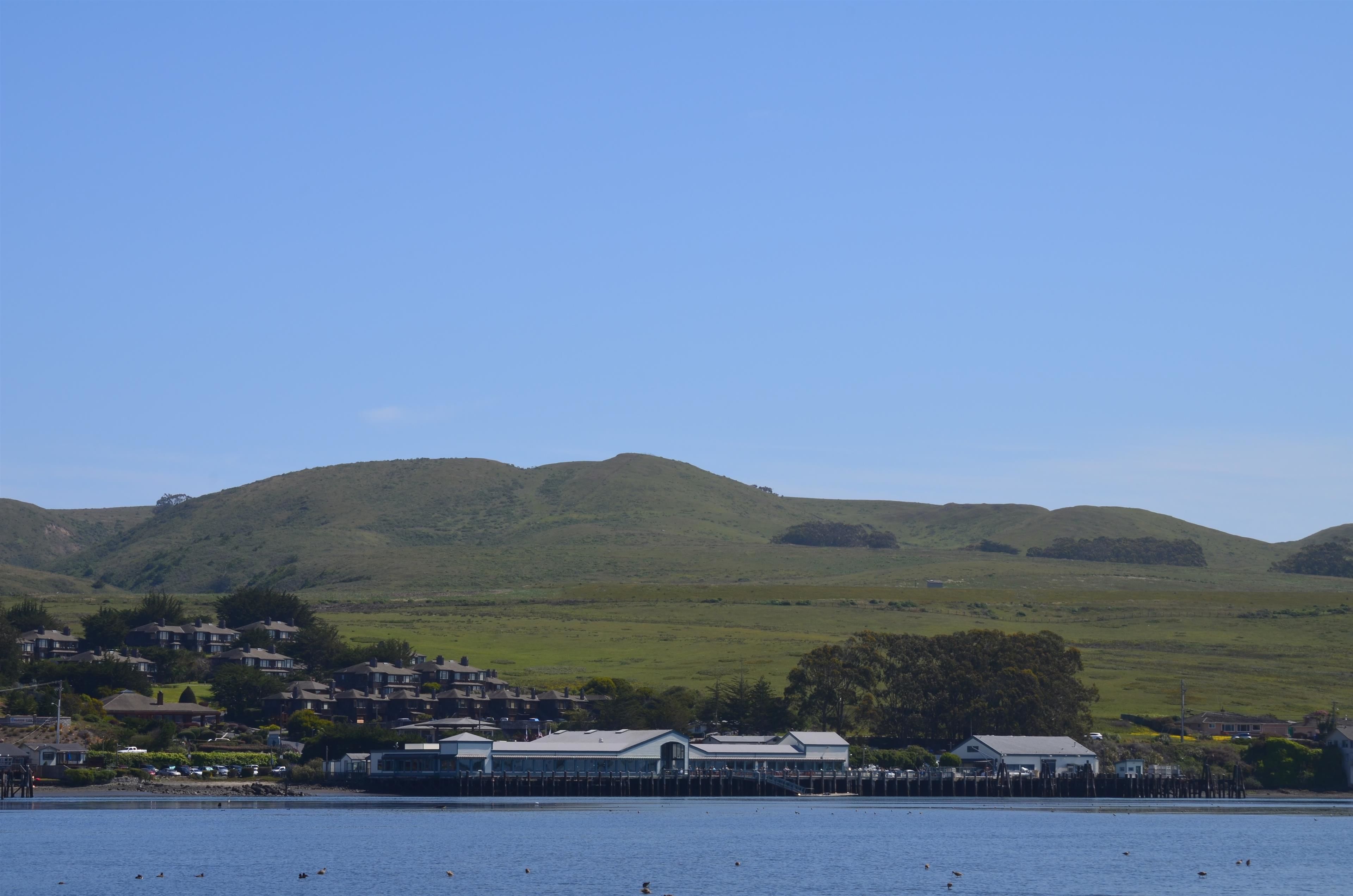 Uma vila costeira com casas numa encosta, um cais com um barco e um céu azul límpido.
