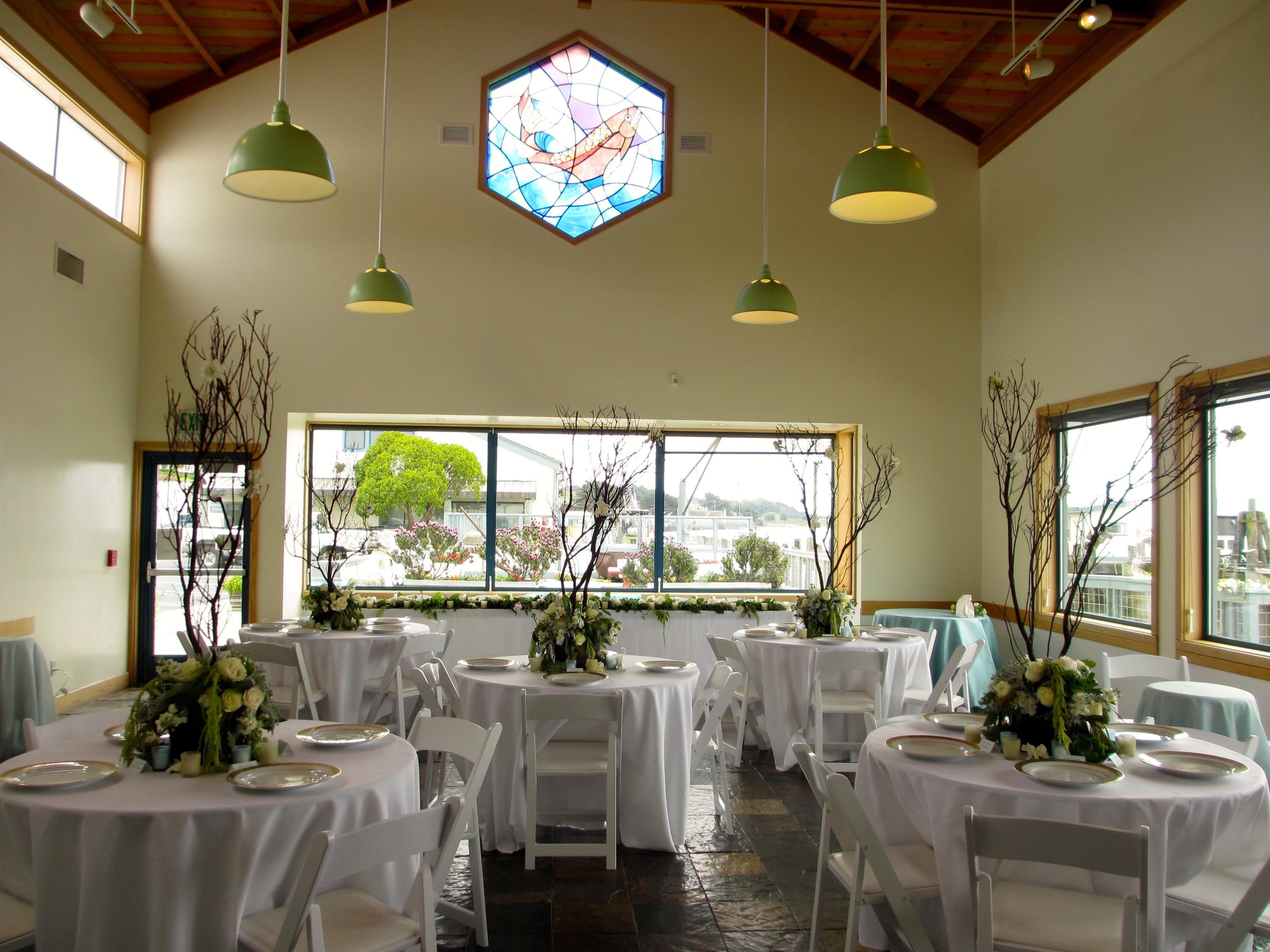 A banquet hall with round tables covered in white tablecloths, white chairs, and green pendant lights. A large stained glass window is visible above the tables.