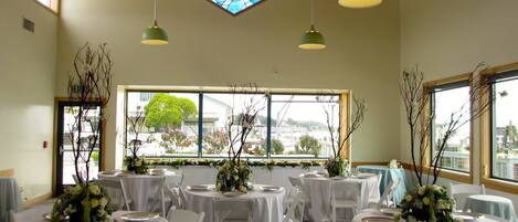 A banquet hall with round tables covered in white tablecloths, white chairs, and green pendant lights. A large stained glass window is visible above the tables.