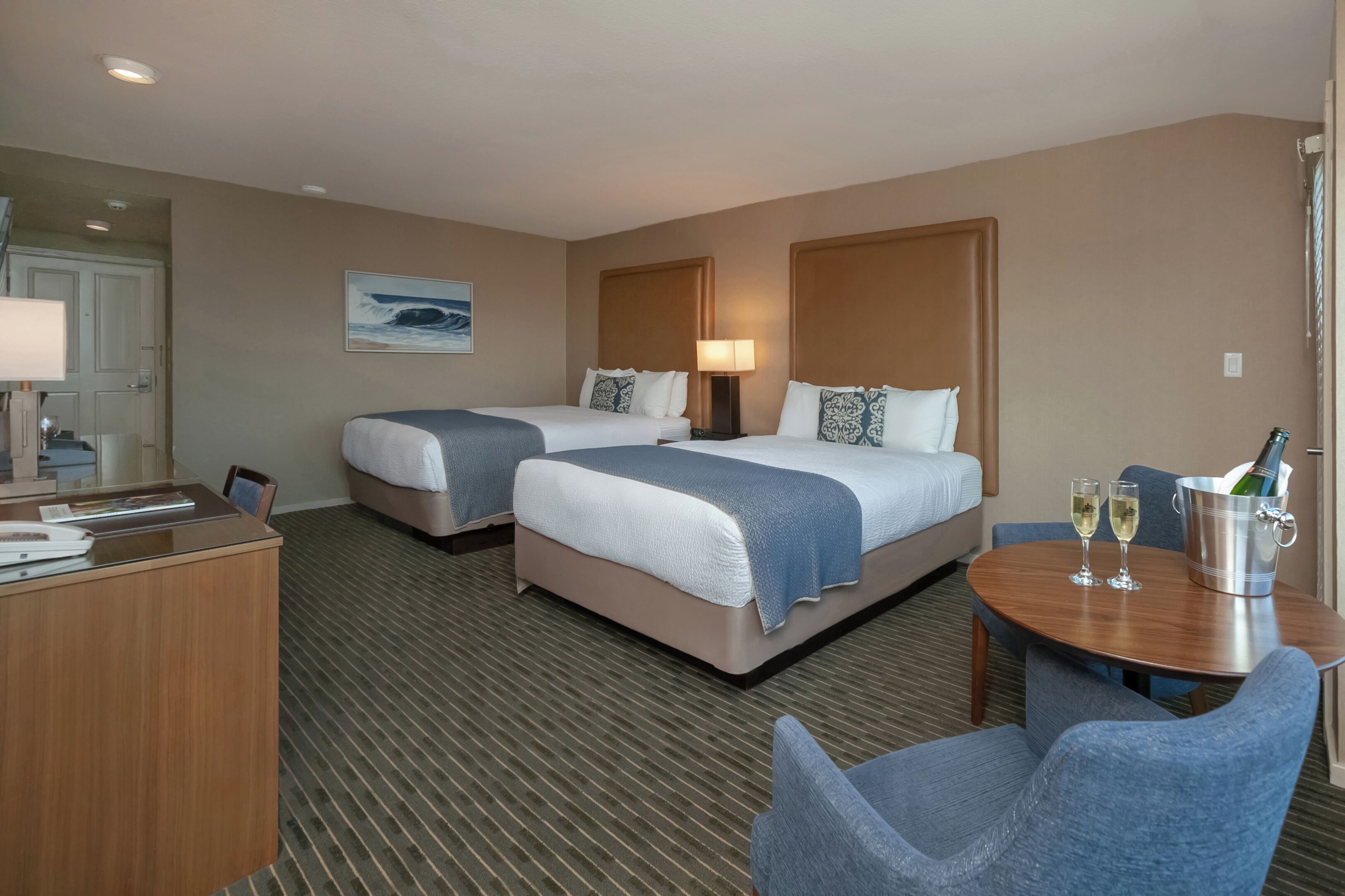 A hotel room with two beds, a desk, a chair, a table with two glasses and a bottle, and a painting on the wall.