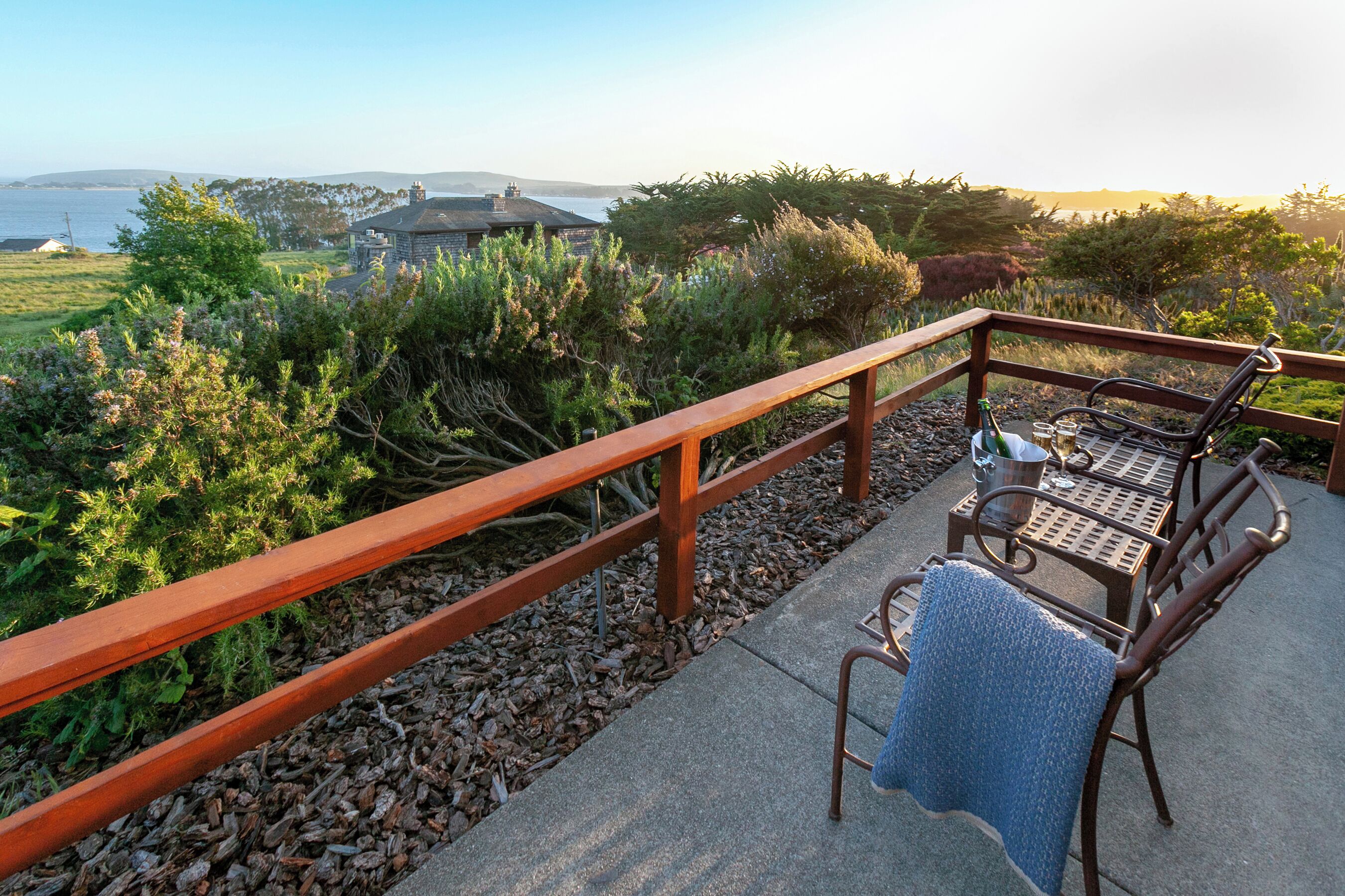A balcony with a wooden railing, a table with chairs, and a view of the sea and hills.