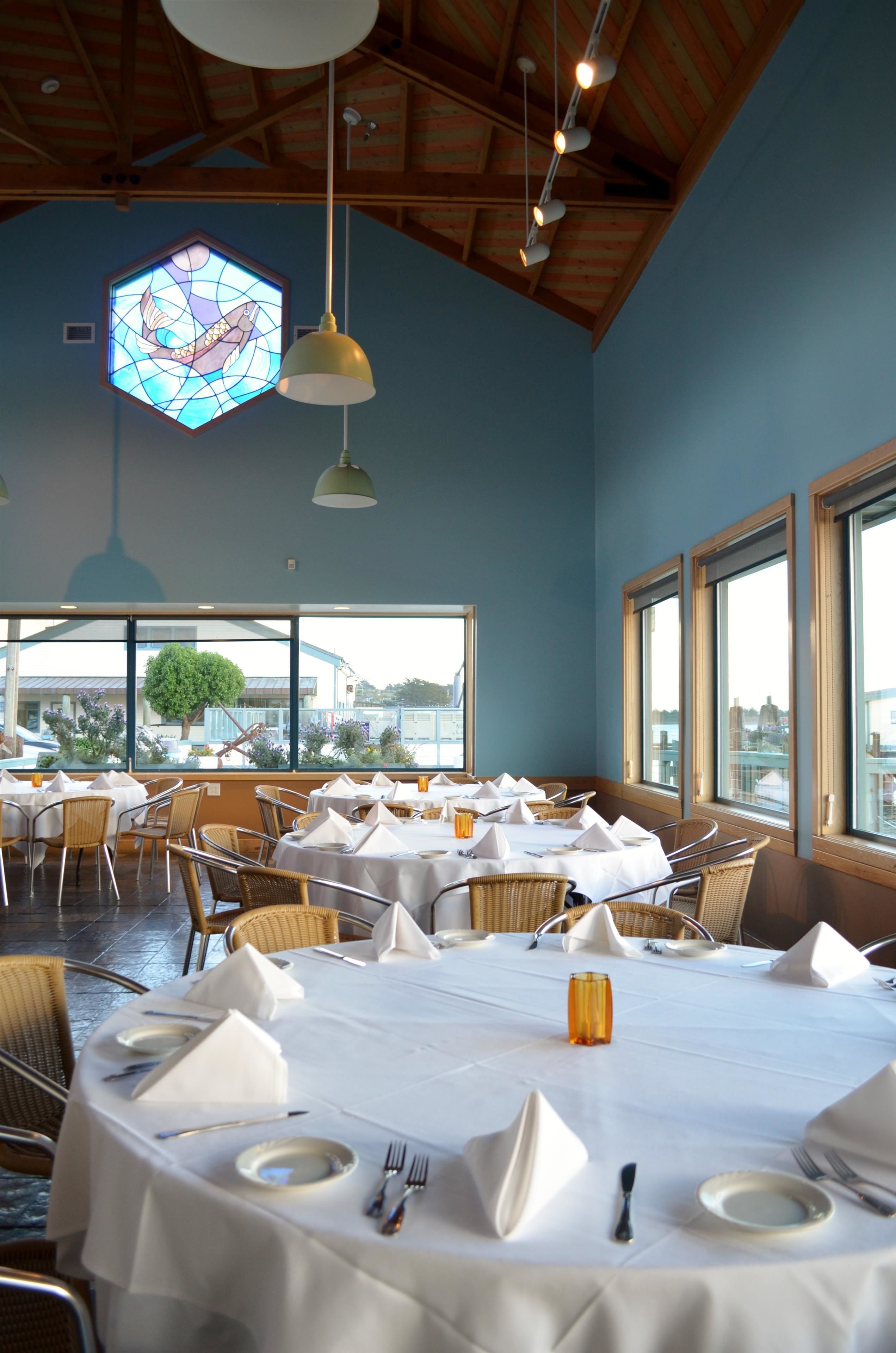 A dining area with round tables set for multiple guests, large windows, and a stained glass window above.