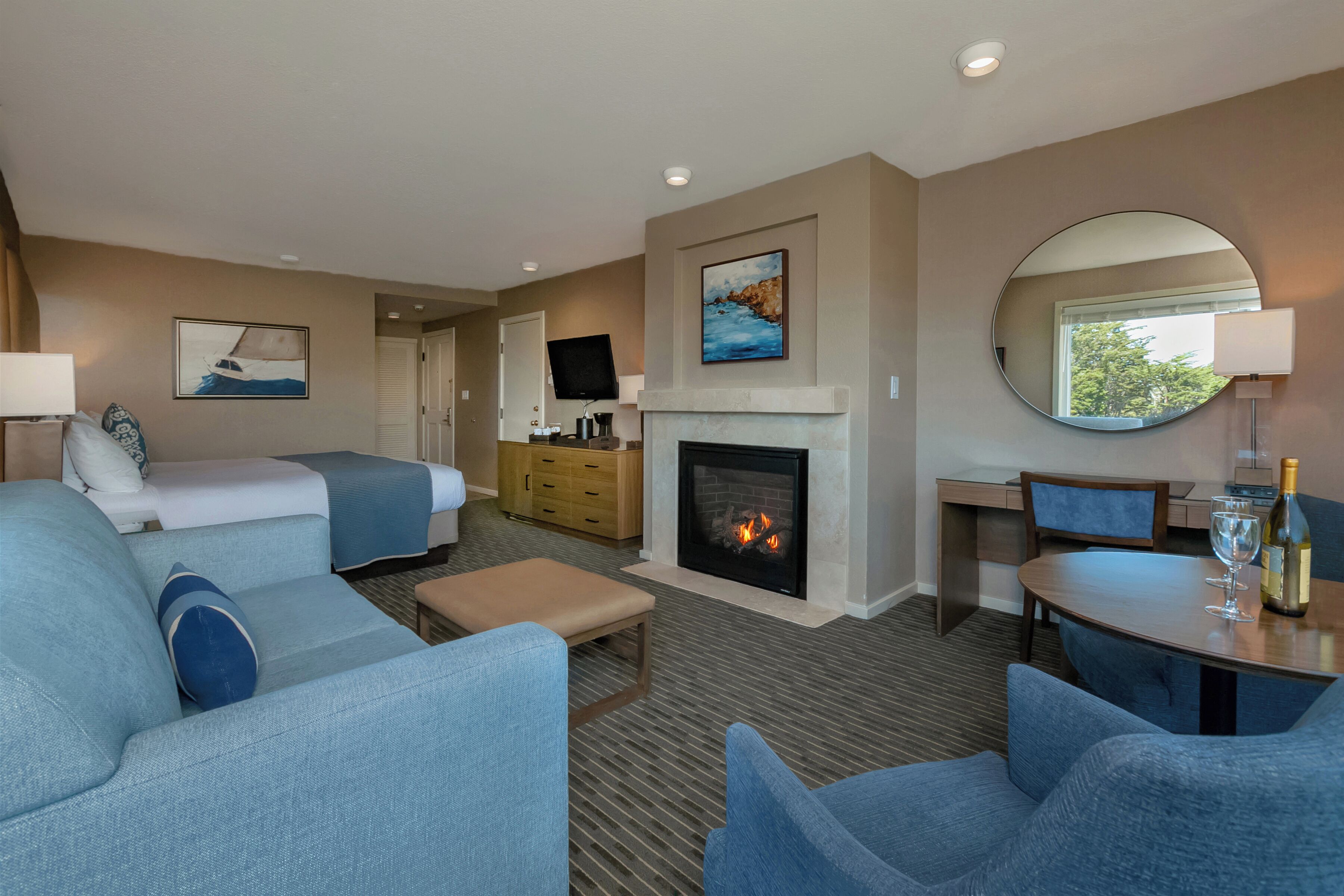 A hotel room with a bed, a sofa, a dining table with a chair, a fireplace, a TV, and a large mirror.
