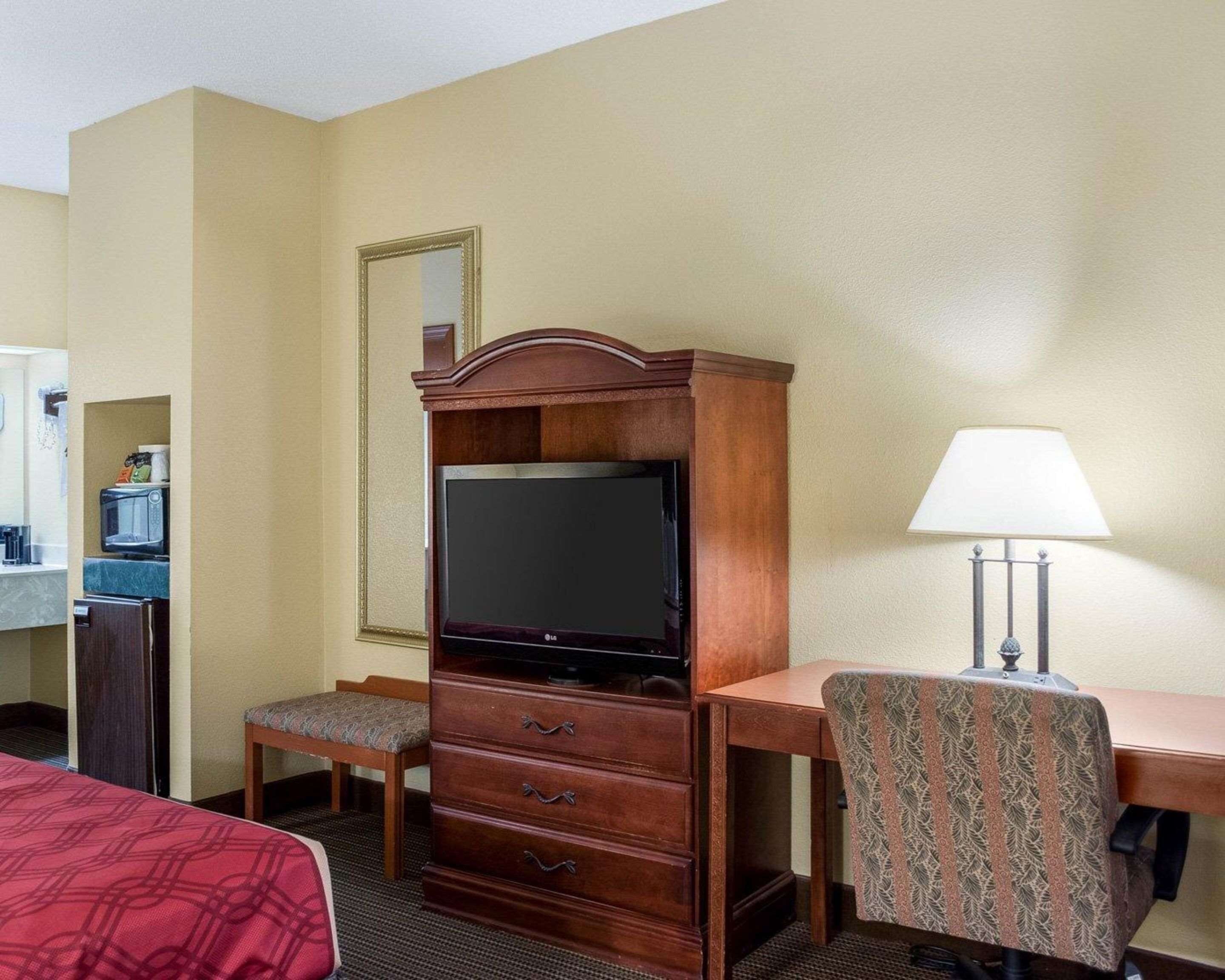 A hotel room with a wooden desk, a television, a patterned armchair, a small bench, and a mirror on the wall.