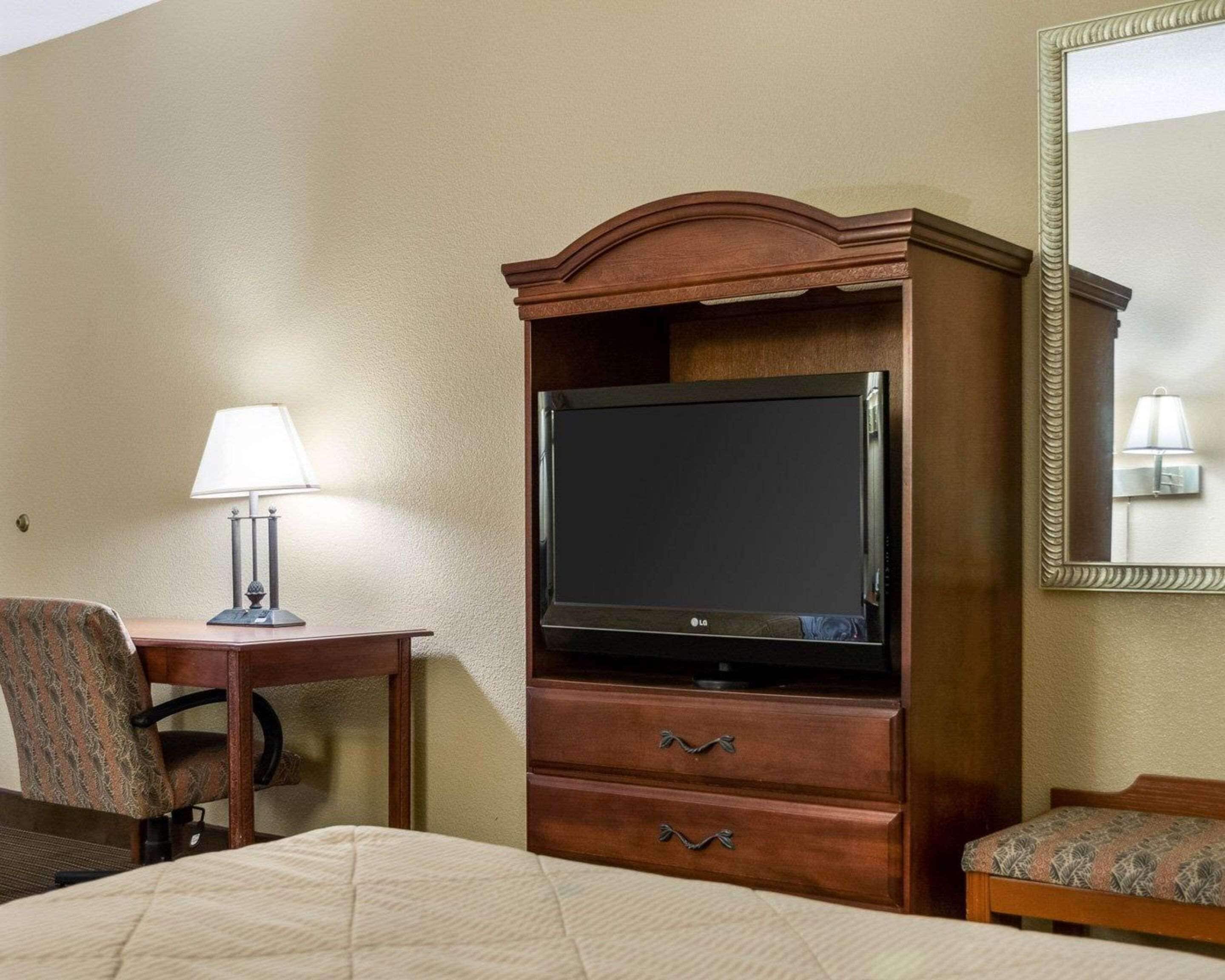 A hotel room with a bed, a wooden dresser with a flat-screen TV, a desk with a lamp, and a chair.