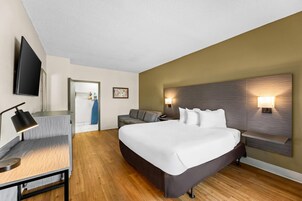 Quarto de hotel com cama, mesa de cabeceira, luminária, televisão e vista para o corredor.