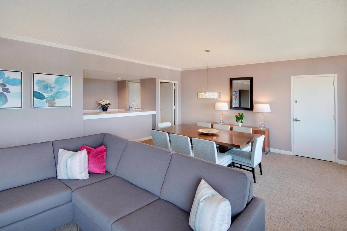 Una sala de estar moderna con un sofá seccional gris, un comedor con mesa y sillas de madera, y un espejo grande sobre la mesa.