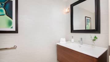 A modern bathroom with a white tiled wall, a wooden vanity, a large mirror, and a framed abstract painting.