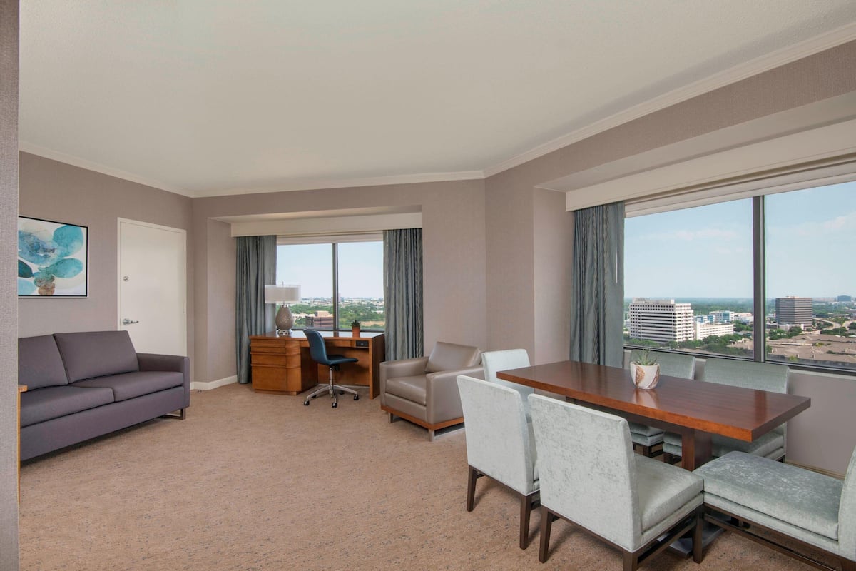 Habitación de hotel con sofá, escritorio, sillas y un amplio ventanal con vista a la ciudad.