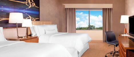 A hotel room with two beds, a desk, a chair, a TV, and a large window with a view of the sky and trees.