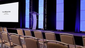 A conference room with a stage, a large screen displaying "The Westin Dallas Park Central", and rows of chairs.