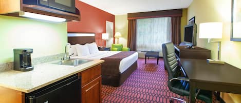 A hotel room with a bed, desk, chair, kitchenette, and a television.